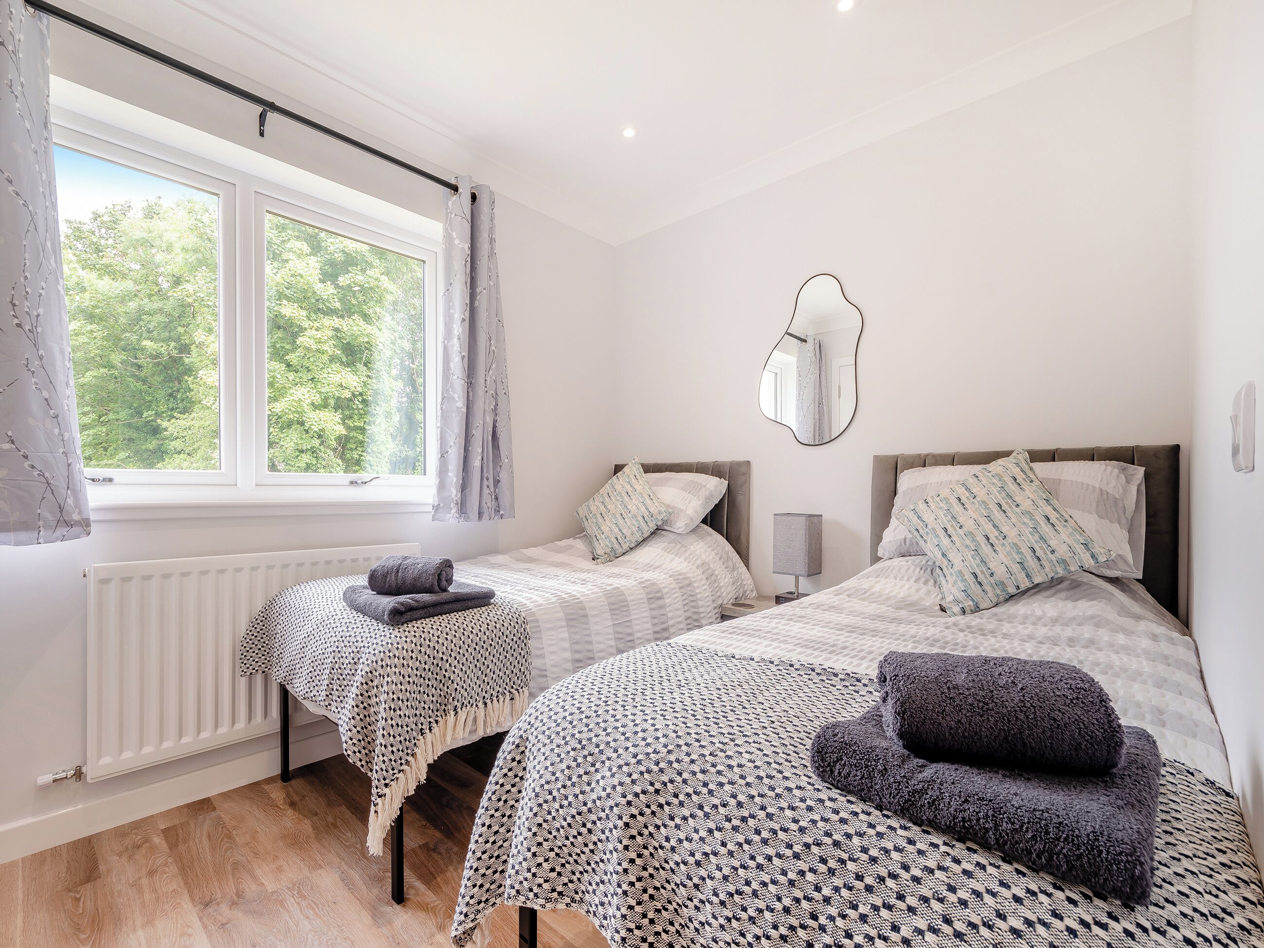A bedroom with two beds, a radiator, a window with curtains, and a mirror on the wall.