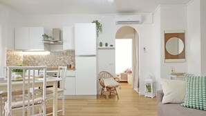A modern kitchen with white cabinetry, a dining area with a white table and chairs, and a living room with a sofa and cushions.