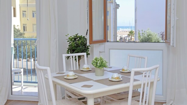 A dining area with a table set for four, white chairs, a potted plant, and a view of the sea through a balcony.