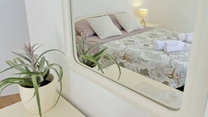 A hotel room with a bed, pillows, a bedside lamp, a potted plant, and a window with a view of another room.
