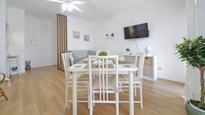 A dining area with a white table and chairs, a ceiling fan, a TV mounted on the wall, and a potted plant.