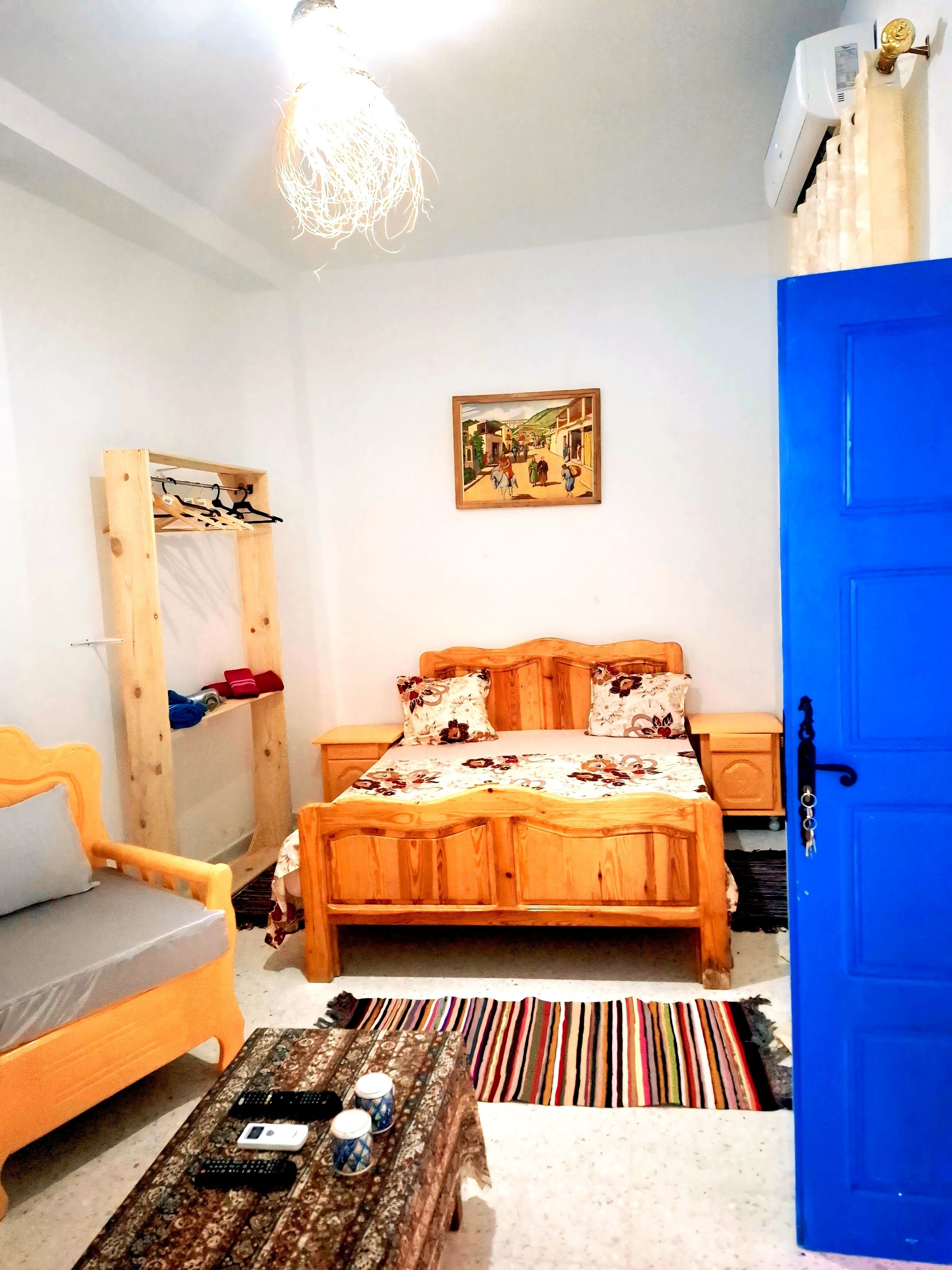 A bedroom with a bed, a chair, a small table with remotes, a colorful rug, and a blue door.