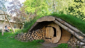 A rustic wooden structure with a thatched roof, surrounded by logs and greenery. - Modern Chalet With Dishwasher, Near the Efteling (Kaatsheuvel)