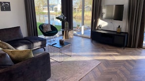 A modern living room with a large window, a sofa, a chair, a coffee table, and a TV. - Modern Chalet With Dishwasher, Near the Efteling (Kaatsheuvel)