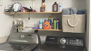 Un'area lavanderia con lavatrice e asciugatrice a carica frontale, mensole sopra con detersivi, uno scaldabagno e un cesto portaoggetti.