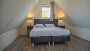 Ein Schlafzimmer mit schrägem Dach, einem Bett mit zwei Kissen, zwei Nachttischen mit Lampen und einem Fenster mit Vorhängen.