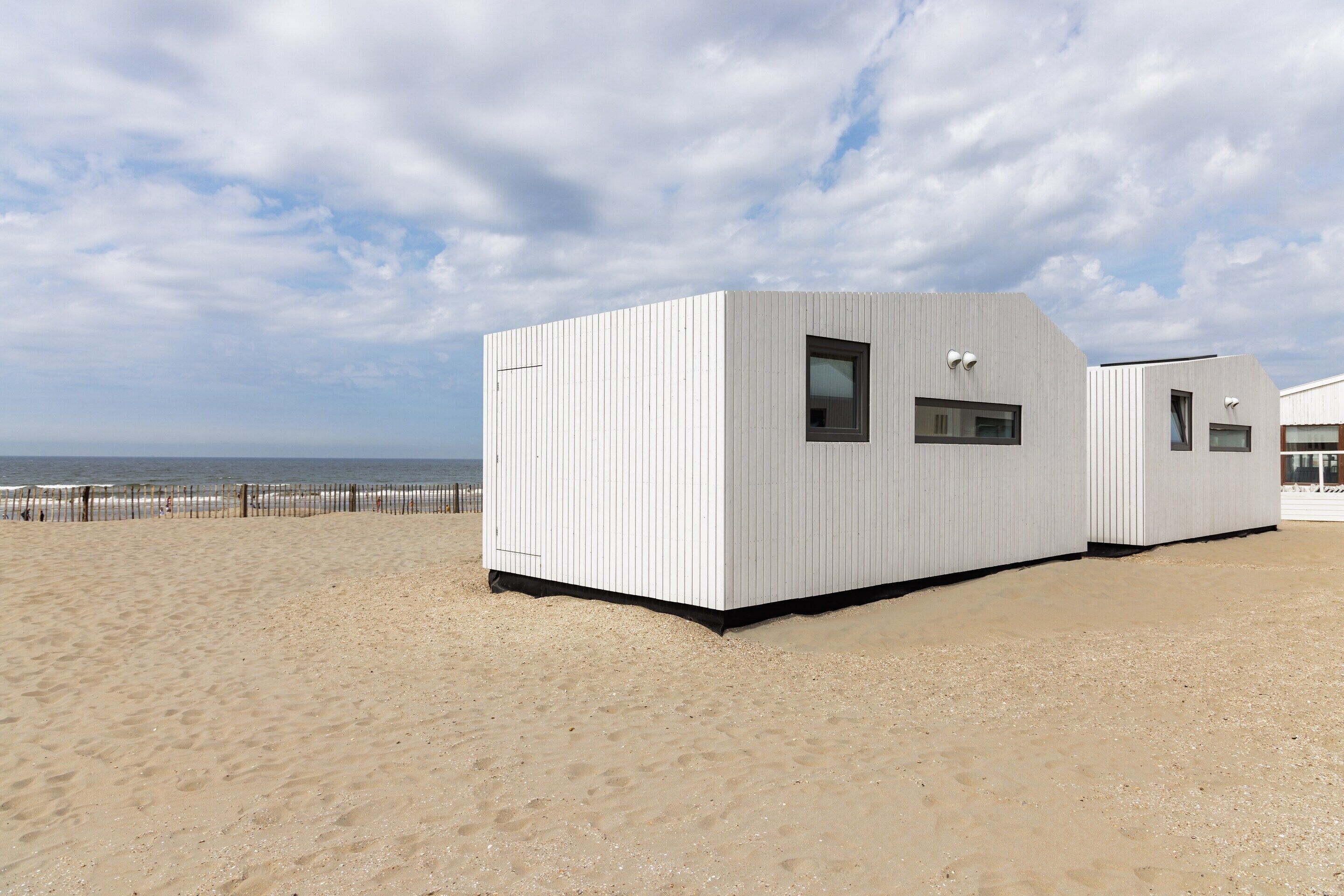 A modern beachside building with a flat roof and large windows, situated on a sandy beach with a view of the ocean.