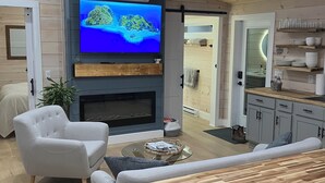 Una sala moderna con un televisor de pantalla plana, una chimenea, un comedor con mesa y sillas de madera, y una cocina con armarios grises y una puerta de vidrio.