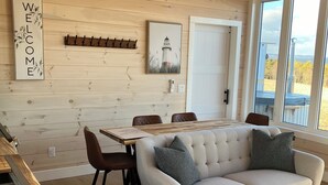 Interior acogedor de una cabaña con techo de madera, una zona de comedor con sofá y sillas blancos, un perchero en la pared y una pintura de un faro.