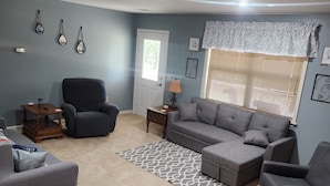 A living room with a grey sofa set, a dark armchair, a patterned rug, and a window with blinds.