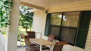 A covered patio with a table and four chairs, a hanging flower arrangement, and a view of a green tree through a window.
