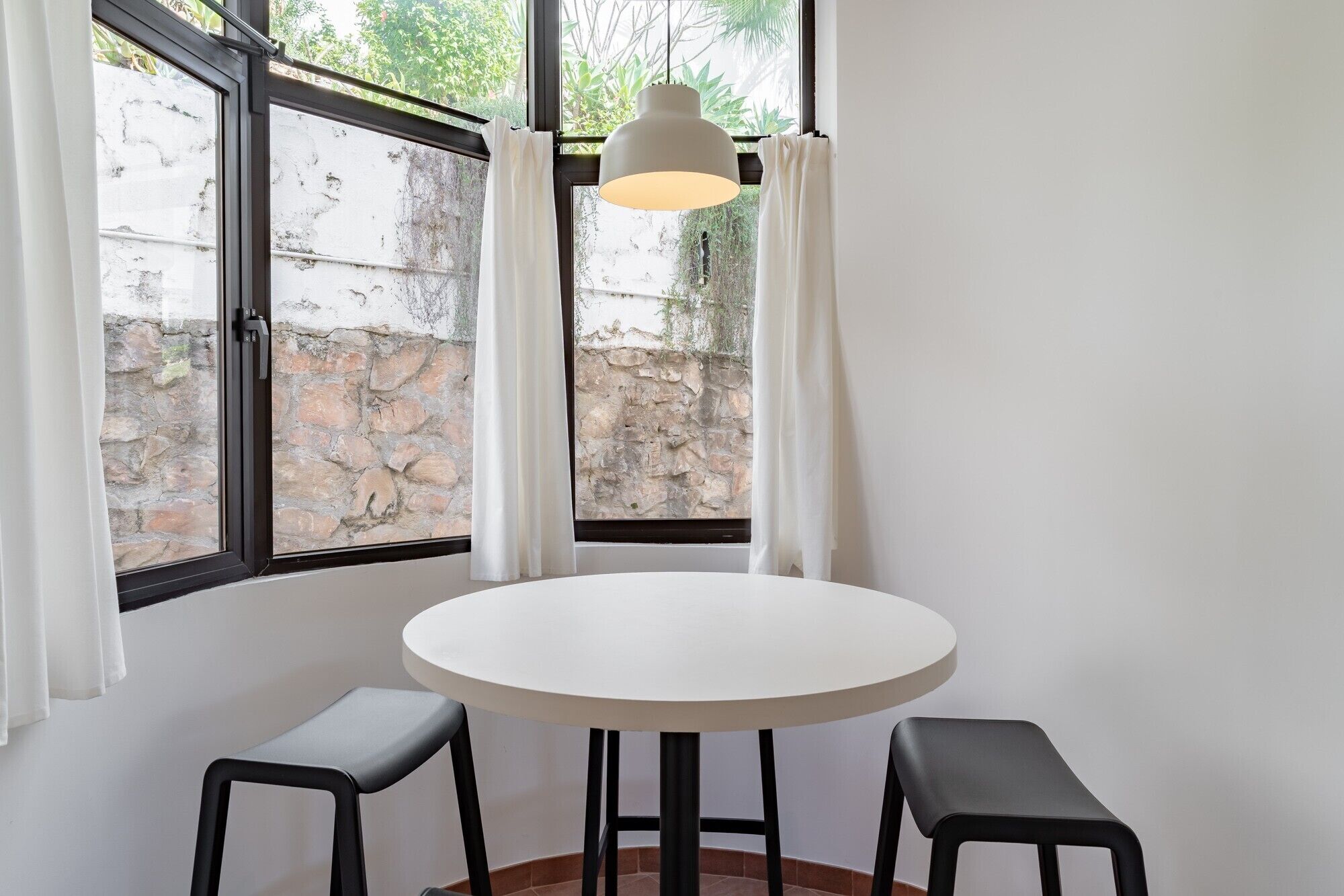 Una mesa redonda con dos taburetes negros, una lámpara colgante y vistas a un muro de piedra a través de amplias ventanas con cortinas blancas.