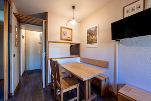 Uma mesa de jantar de madeira com bancos, uma TV fixada na parede, quadros emoldurados e uma porta que dá para outro quarto.