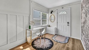 Una habitación con una puerta blanca, un escritorio con silla, un armario pequeño blanco y un espejo redondo en la pared.