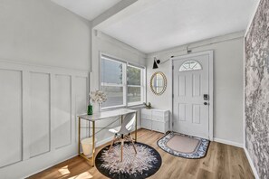 Una habitación con una puerta blanca, un escritorio con silla, un armario pequeño blanco y un espejo redondo en la pared.