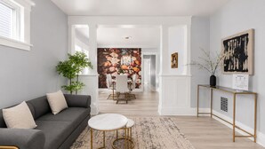 Un salon moderne avec un canapé gris, une table basse ronde, un coin repas avec une table et des chaises blanches, et un couloir menant à la cuisine.