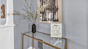 Une pièce avec une cheminée blanche, une œuvre d’art encadrée, un vase avec des branches et un panneau d’accueil.