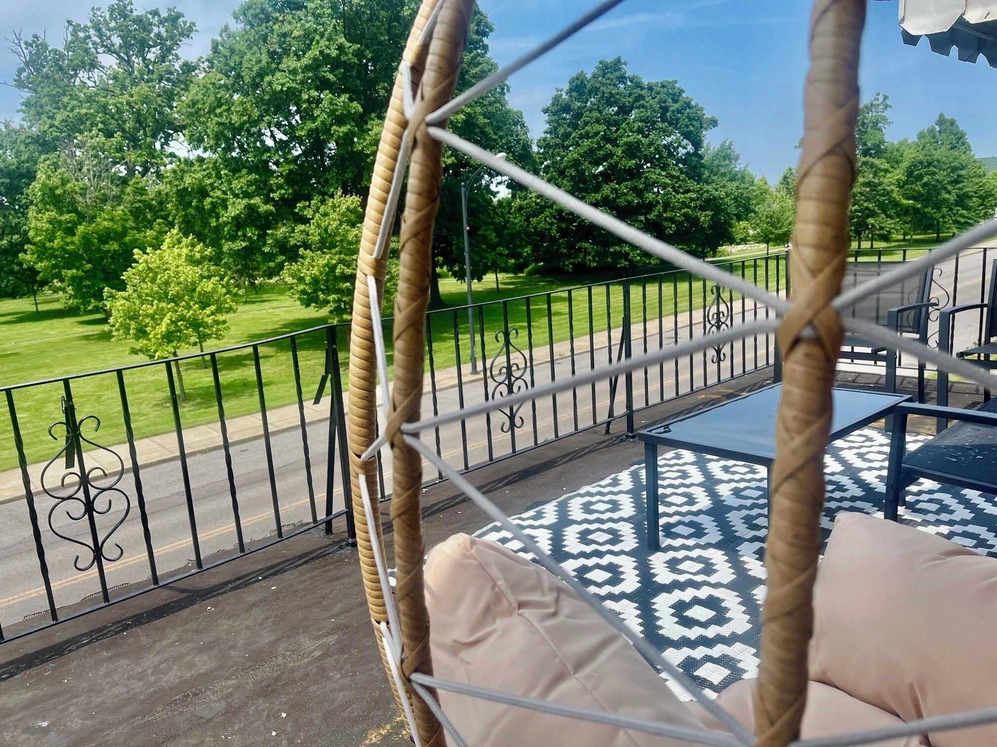 Ein Korbschaukelstuhl mit einem gemusterten Boden, einem Metallgeländer und Blick auf einen grünen Park mit Bäumen und einer Straße.