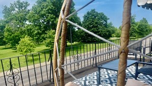 Een rieten hangstoel met een patroon in de vloer, een metalen leuning en uitzicht op een groen park met bomen en een weg.