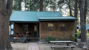 Un rifugio rustico con tetto in lamiera verde, un portico con sedie di legno e un tavolo da picnic.