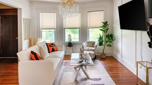 A living room with a white sofa, a glass coffee table, a chandelier, and a large TV on the wall. - Niagara RiversideView Balcony GameRoom WiFi Free Parking King bed Crib sleep 9 (Buffalo)