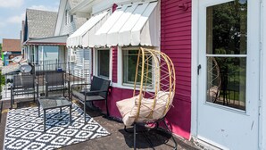 A patio with outdoor furniture, including a swing chair, a table, and chairs, against a house with a white awning and a red exterior wall. - Niagara RiversideView Balcony GameRoom WiFi Free Parking King bed Crib sleep 9 (Buffalo)