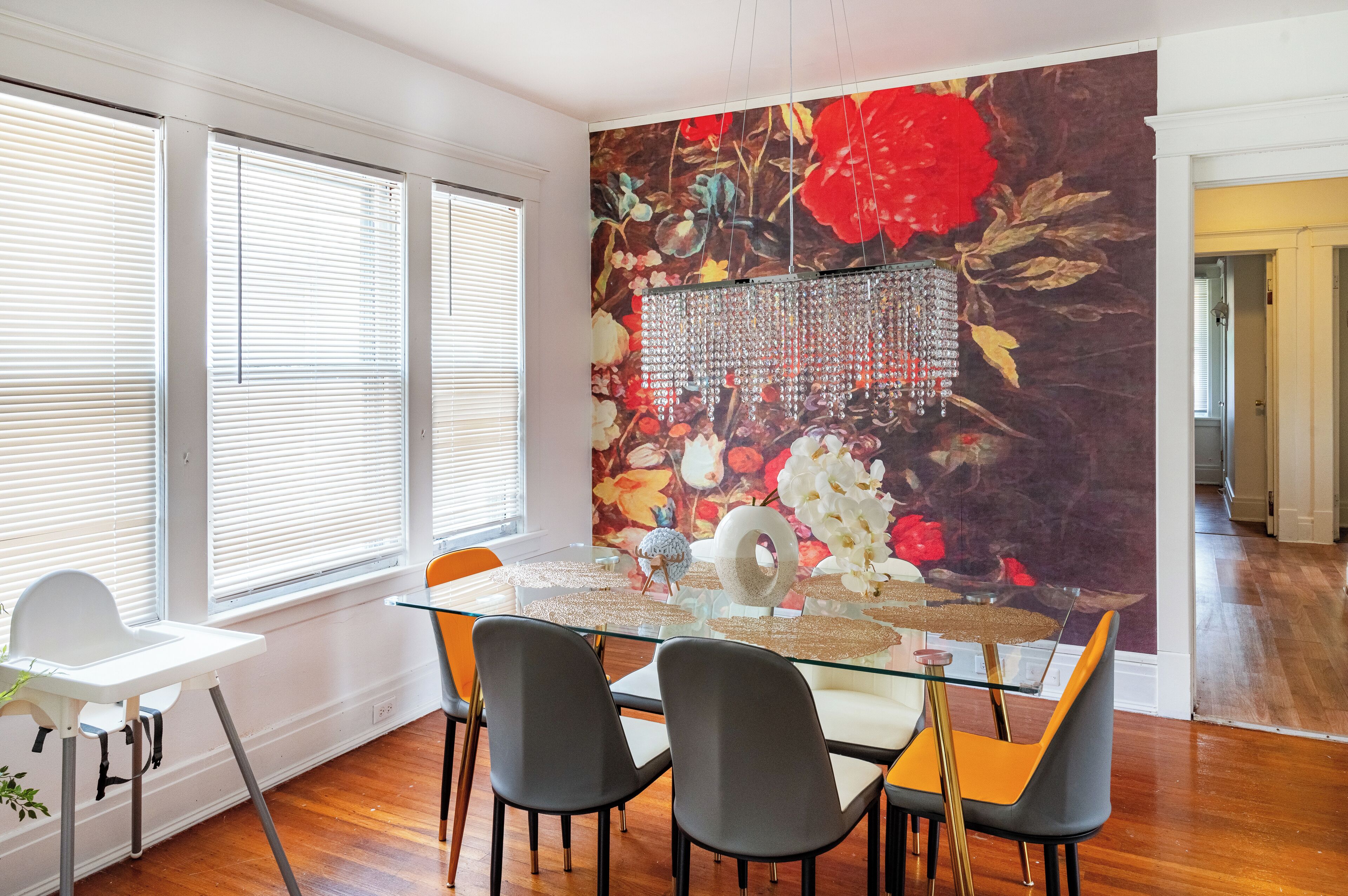 Un coin repas avec une table en verre, des chaises noires et orange, une chaise blanche et un mur décoratif orné d’une fresque florale.