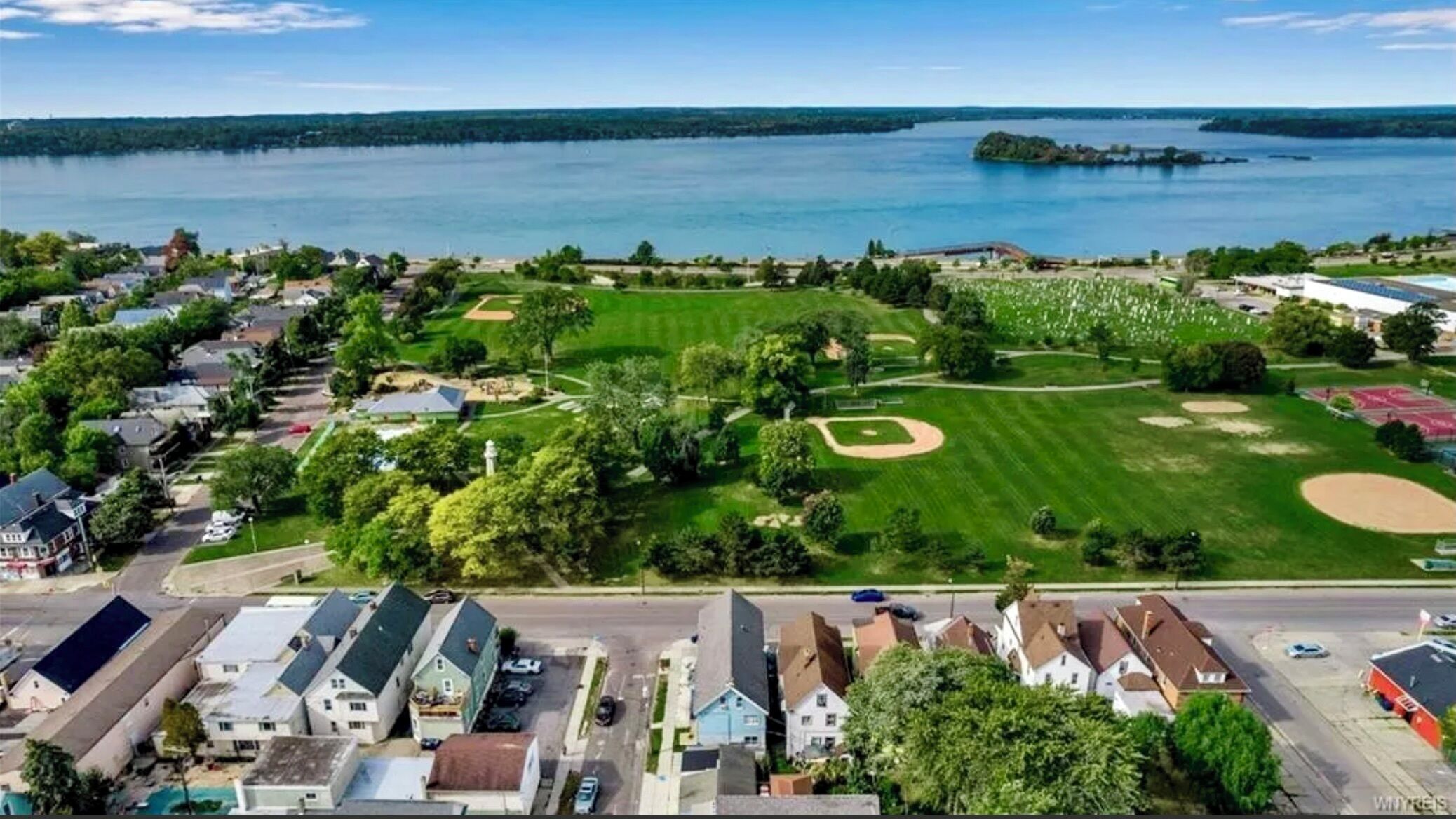 A residential area with houses, a park with a baseball field, and a large body of water in the background.