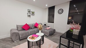 A modern living room with a grey sofa, pink and grey cushions, a round coffee table with a magazine, and a black TV stand. - Luxury One Bedroom Apartment in Berkshire  (Reading)