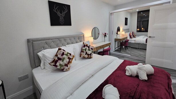 A modern hotel room with a large bed, a bedside table with a lamp, a mirror, and a wall-mounted artwork. - Luxury One Bedroom Apartment in Berkshire  (Reading)