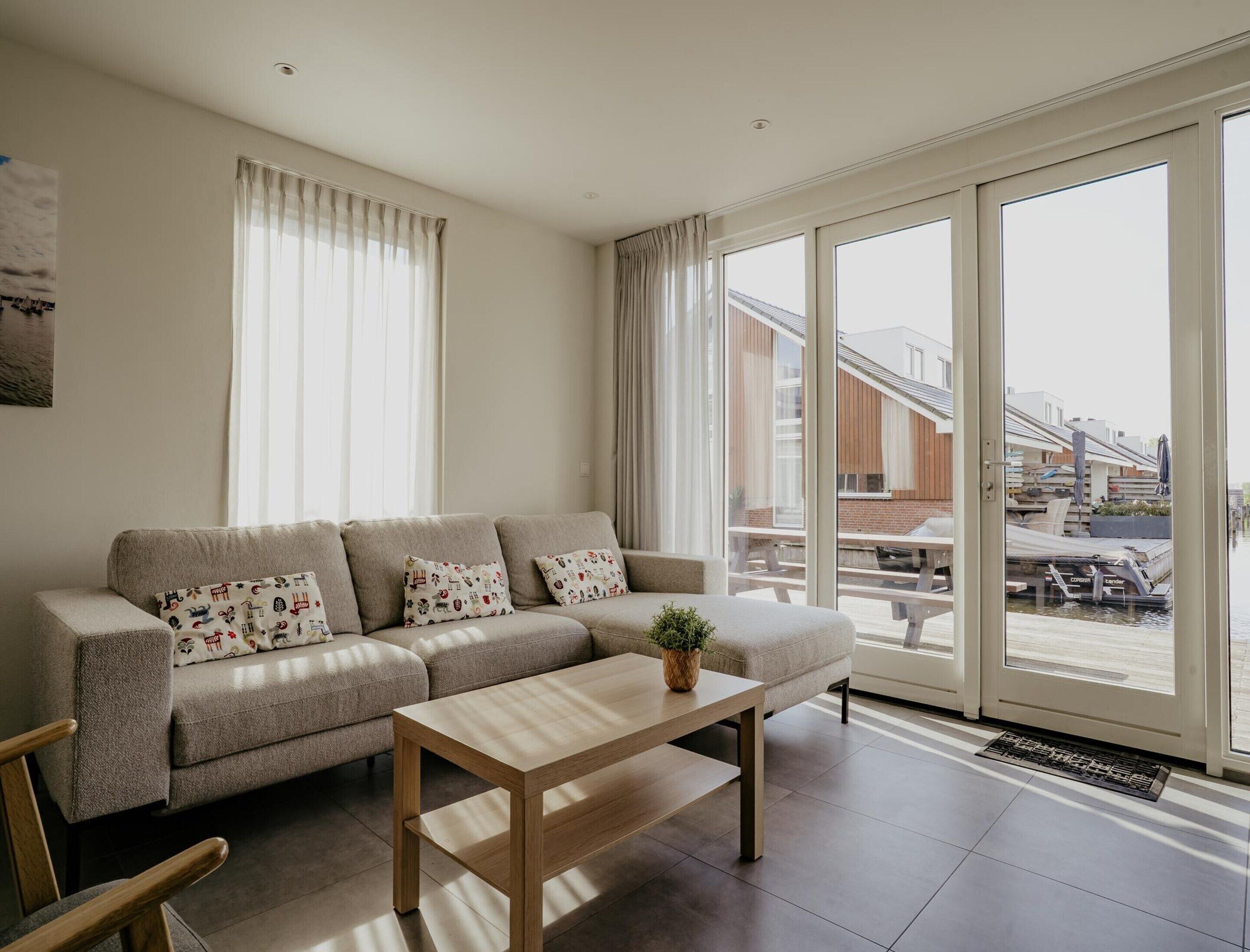 A modern living room with a sofa, a wooden coffee table, and large windows offering a view of a building with a sloped roof.