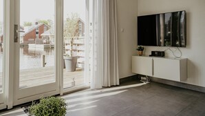 A modern living room with a large sliding glass door leading to a balcony, a flat-screen TV mounted on the wall, and a minimalist white cabinet. - Waterside Escape With Easy Access (Uitgeest)