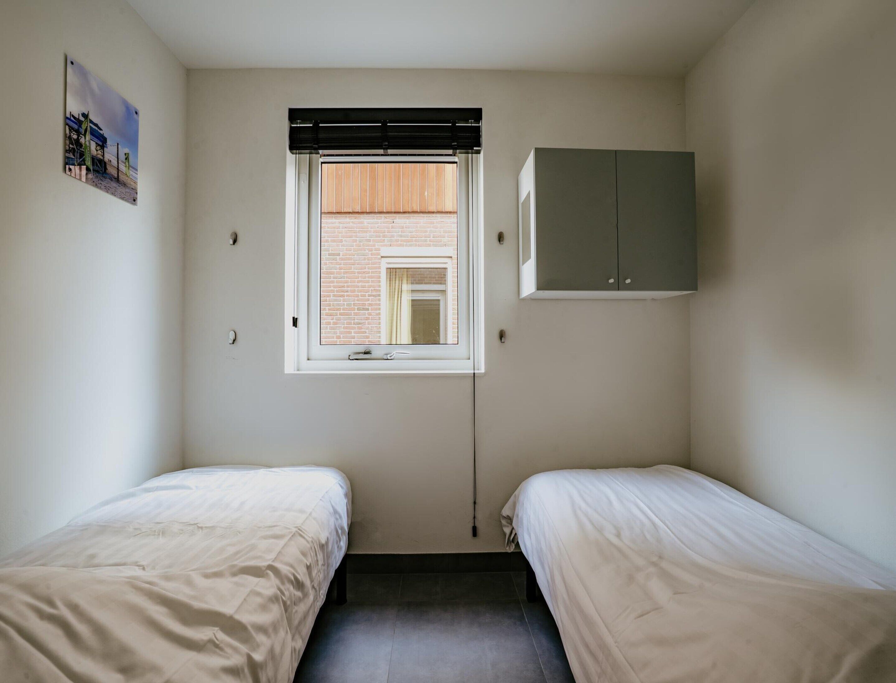 A small room with two single beds, a window with blinds, a wall-mounted cabinet, and a picture on the wall.