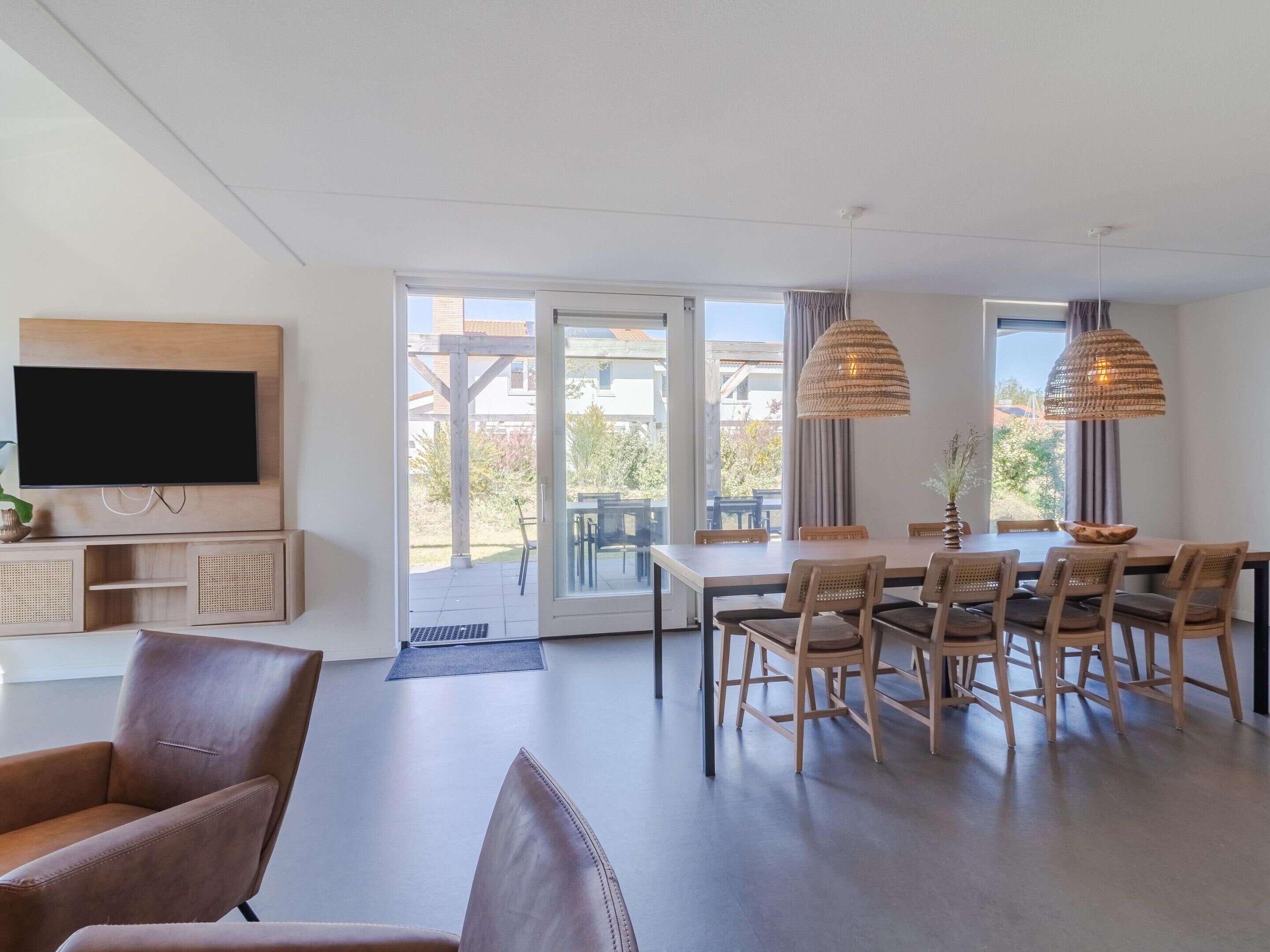 A modern living room with a flat-screen TV, wooden furniture, and a dining area with a view.
