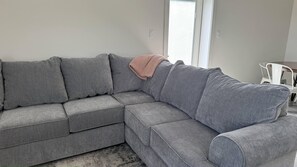 Harmaa kulmasohva, jonka päällä on vaaleanpunainen huopa, valkoinen tuoli ja ikkuna, jossa on sälekaihtimet.