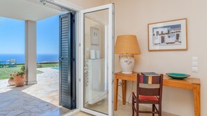 Une porte-fenêtre coulissante donne sur une terrasse offrant une vue sur la mer. On y trouve une table avec une lampe, une chaise et un tableau encadré accroché au mur.