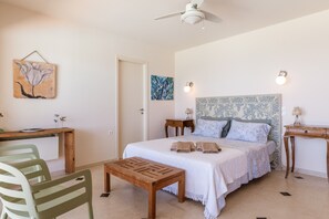 A bedroom with a bed, bedside tables, a desk, chairs, a ceiling fan, and artwork on the walls.