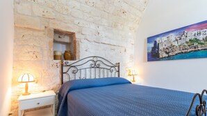 Une chambre avec un mur en briques blanches, un cadre de lit en métal, une couvre-lit bleue, une table de chevet avec une lampe et un tableau représentant une ville côtière.