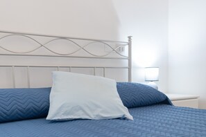 Un lit avec une tête de lit blanche et une literie bleue, une table de chevet avec une lampe, et un mur blanc.