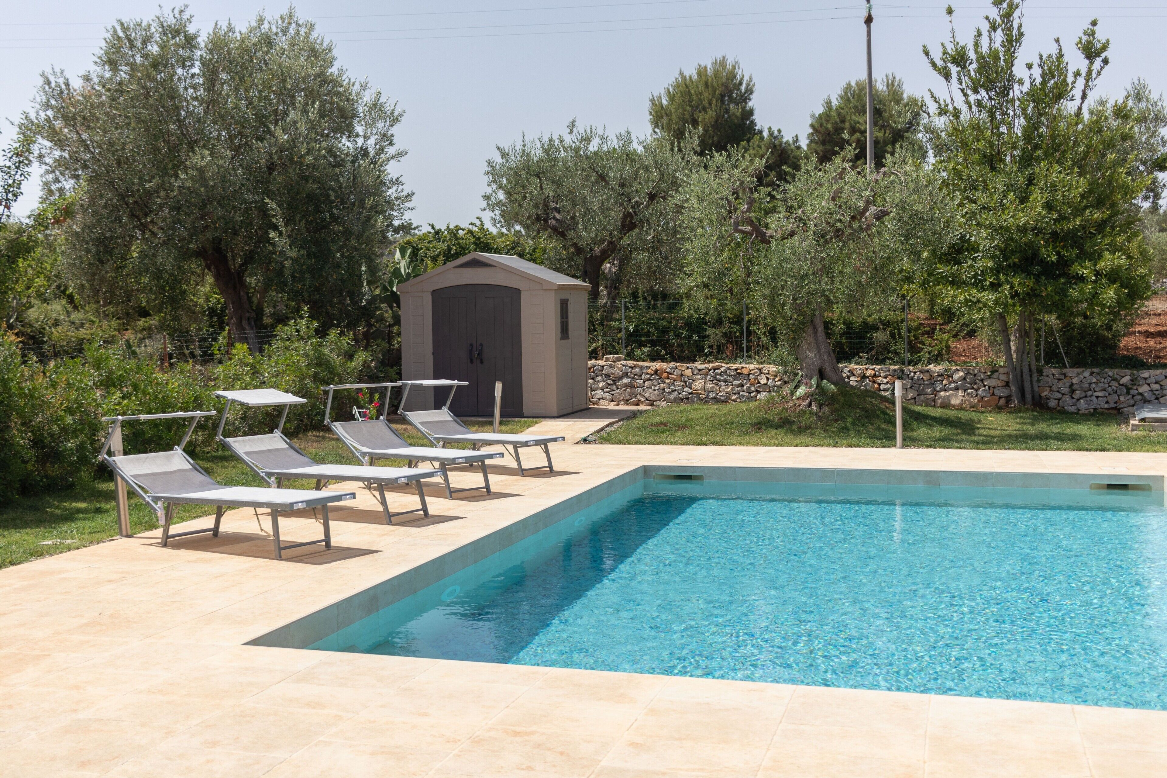 A rectangular swimming pool with clear blue water, surrounded by a tiled deck. There are four lounge chairs aligned along the pool's edge.