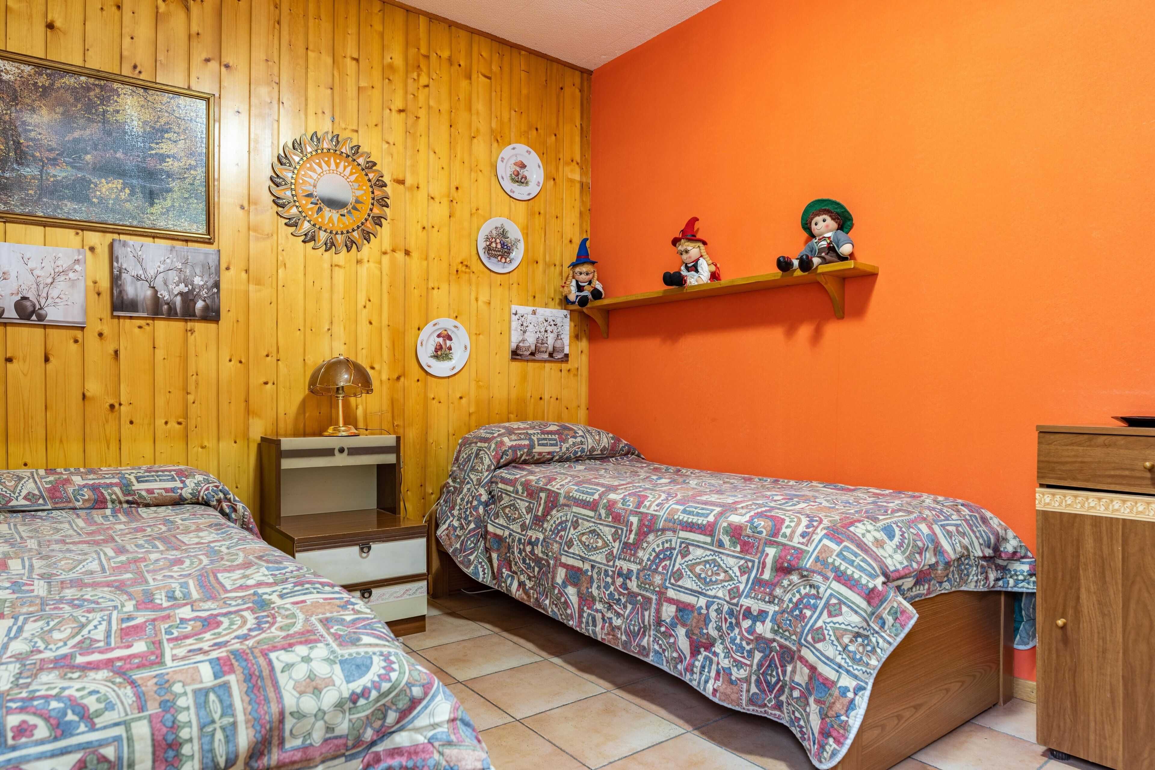 A room with two beds, wooden walls, a shelf with figurines, and framed pictures.