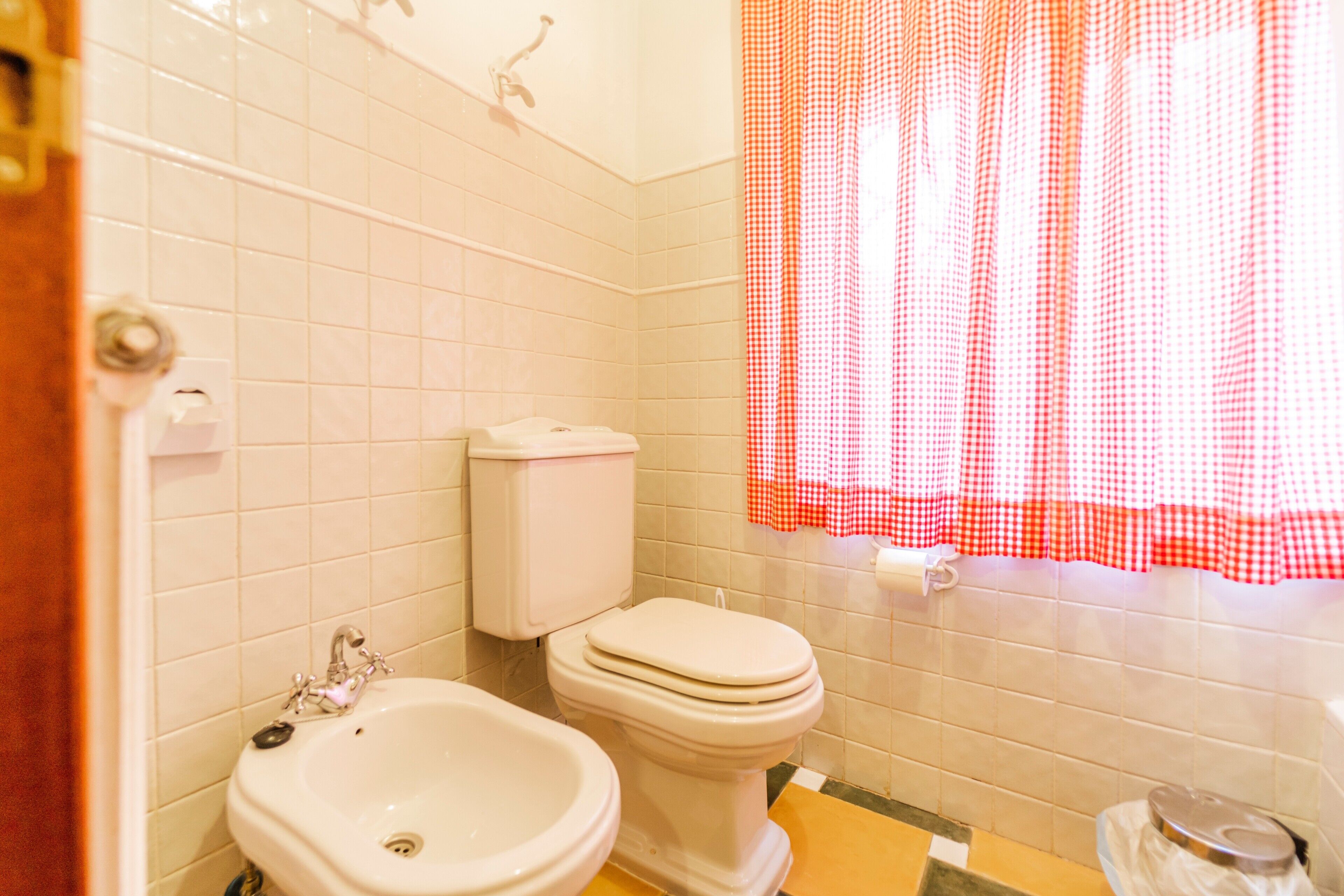 Un baño con azulejos blancos, un lavamanos, un inodoro y una cortina de ducha a cuadros roja y blanca.