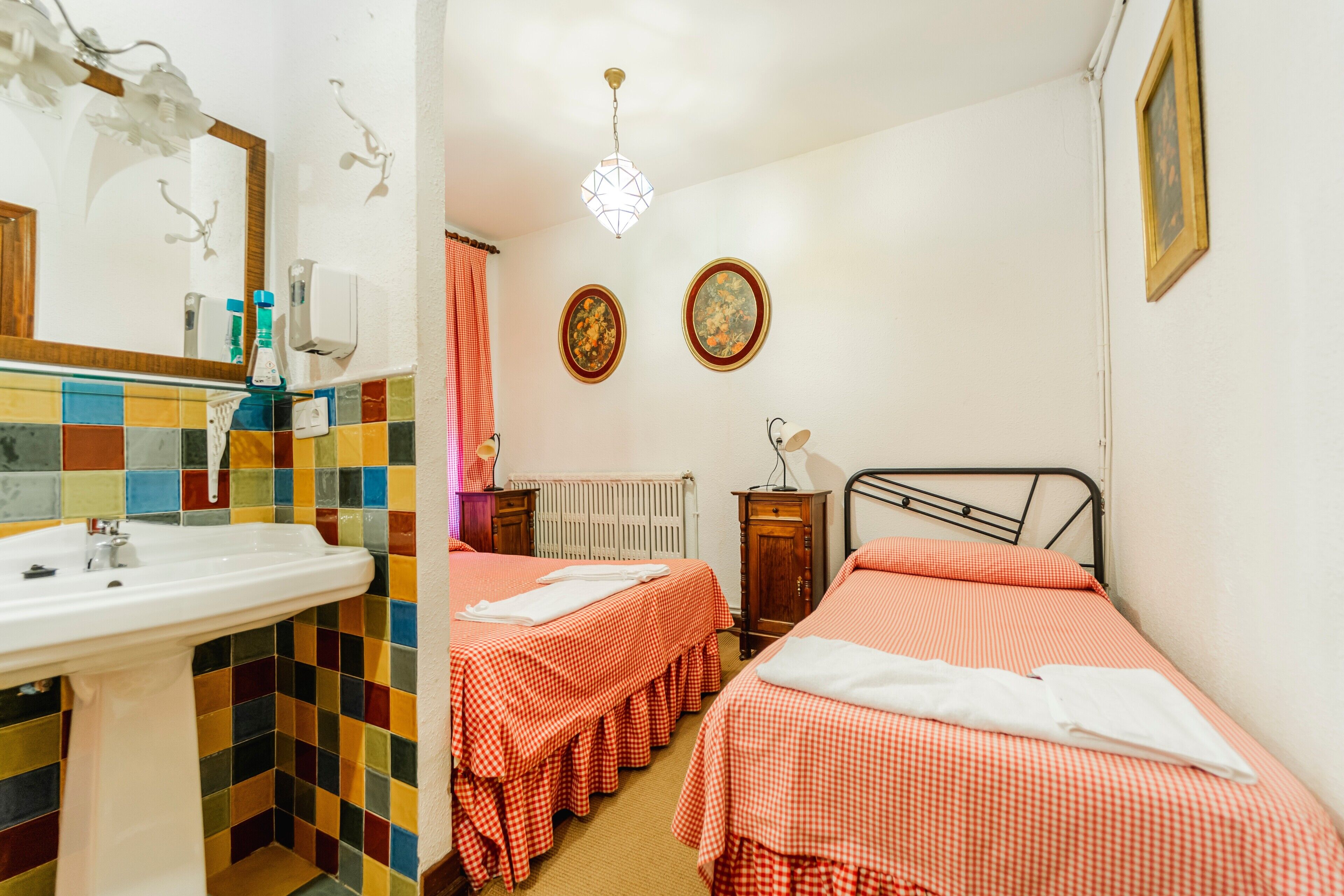 Una habitación con pared de azulejos, un lavabo blanco, dos camas con ropa de cama a cuadros, un radiador y dos cuadros enmarcados en la pared.