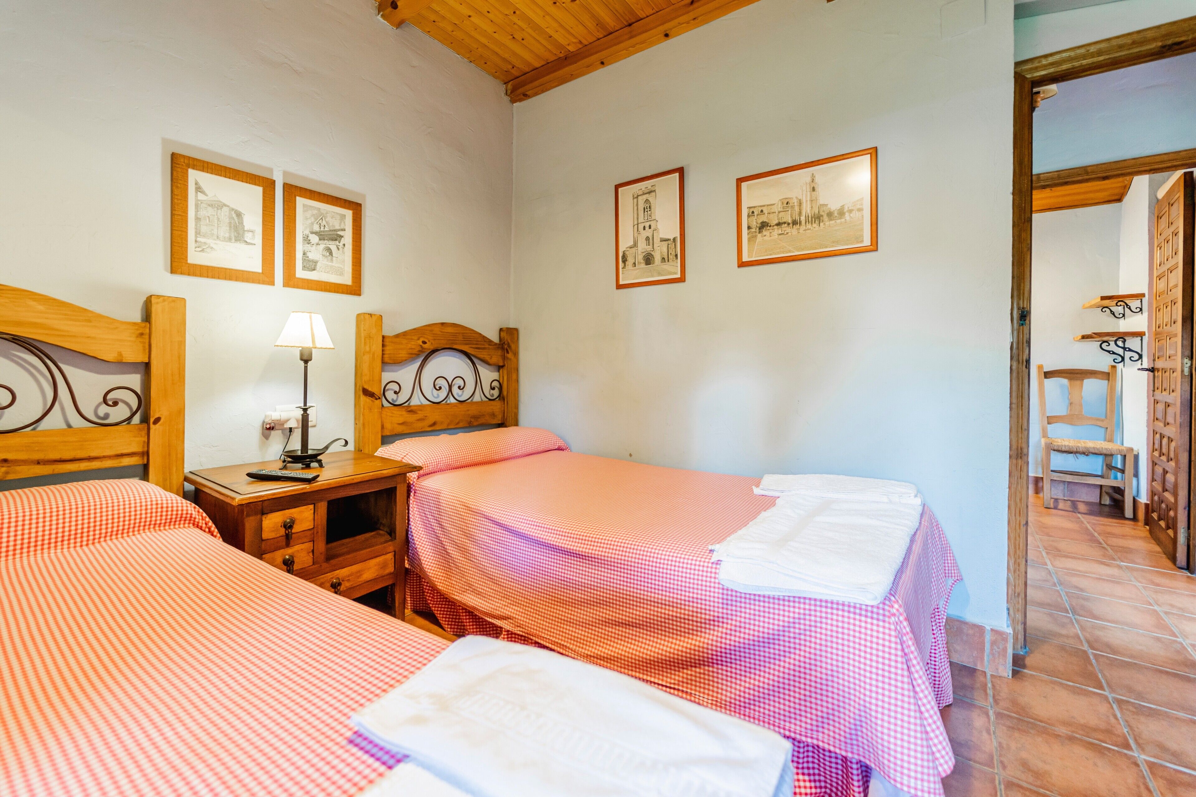 Habitación de hotel con tres camas, cabeceros de madera, una mesita de noche con lámpara y cuadros enmarcados en la pared.