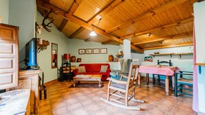 Un interno rustico con soffitto in legno, un divano rosso, una poltrona a dondolo, un tavolo da pranzo con tovaglia a quadretti e una zona cucina sullo sfondo.
