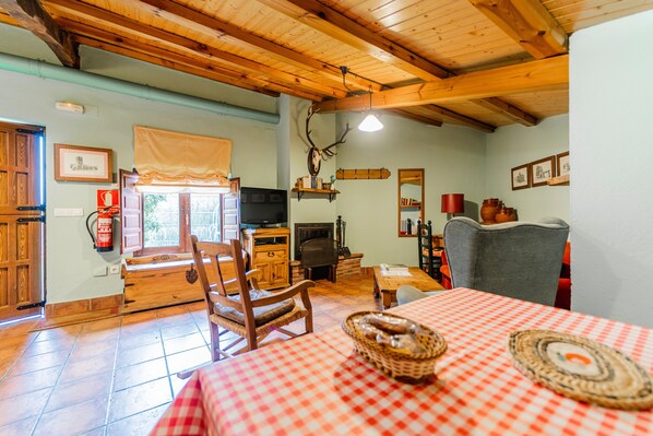 A cozy room with a wooden ceiling, a fireplace, a television, a dining table with a checkered tablecloth, and a rustic decor including a deer head.