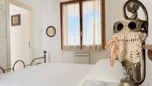 Uma cama com estrutura de metal, uma janela com cortinas, um radiador e um espelho fixado na parede.