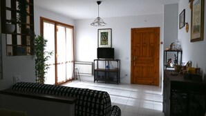 Uma sala de estar com um sofá, uma televisão sobre um suporte, uma porta de madeira e uma janela grande.