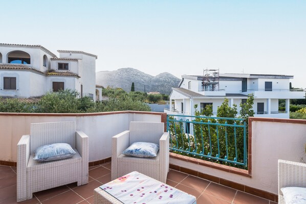 Una terraza con mobiliario de mimbre, una mesa blanca y vistas a edificios y montañas.