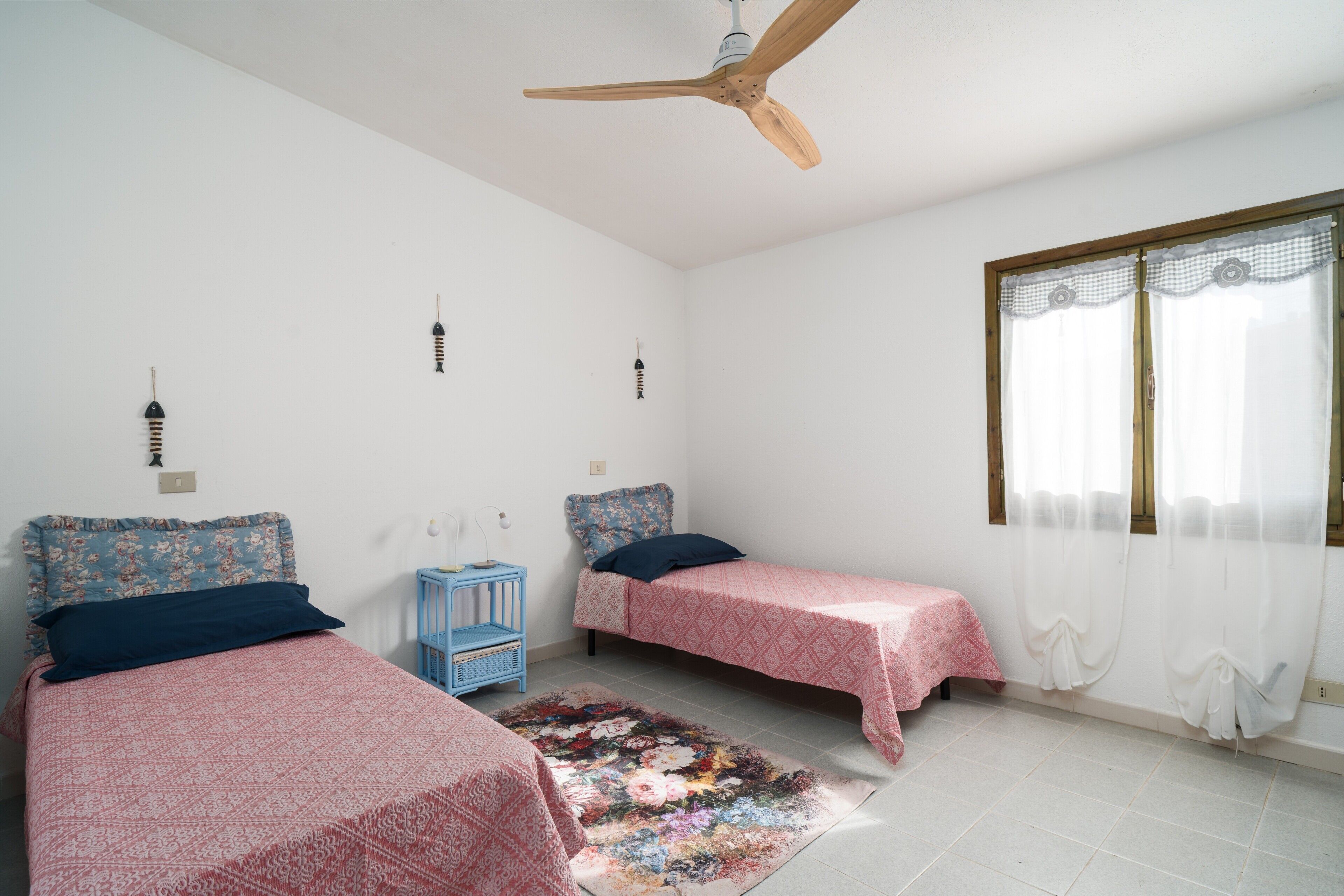 Un dormitorio con dos camas individuales, un ventilador de techo, una ventana con cortinas y una mesita de noche azul pequeña.